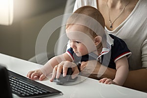 Portrait of cute little baby boy sitting on mothers lap and reaching for computer keyboard