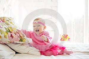 Portrait of a cute little baby girl with a princess crown on the head, in pink dress laughing and lying on the bed