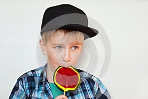 A portrait of a cute little blond boy in black cap and checked shirt eating a bright lollipop isolated over white background looki