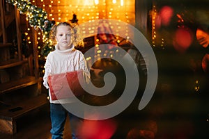 Portrait of cute little blonde curly child girl holding red gift box with Christmas present standing in cozy living room