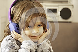 Portrait of cute little blonde girl in soft sweatshirt playing with wireless headphones, listening relaxing music at home. The