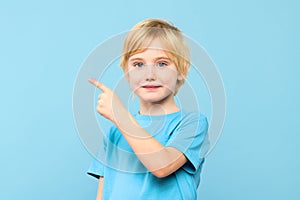 Portrait of a cute little boy with blond hair smiling and pointing to empty place on pastel blue background.