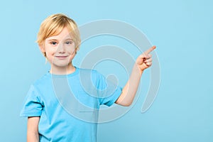 Portrait of a cute little boy with blond hair smiling and pointing to empty place on pastel blue background.