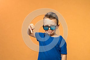 Portrait of a cute little boy giving thumbs down sign with sad expression on orange background.