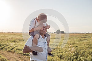 Portrait of cute little boy  and her handsome grandpa looking at camera and smiling on meadow. kid is sitting pickaback