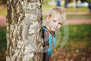 Portrait Cute little boy in a knitted sweater is playing behind a tree in autumn park, play at hide-and-seek