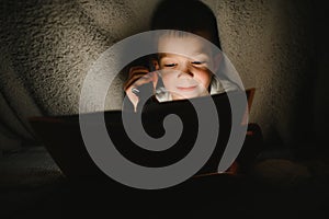 Portrait of cute little boy reading in bed with flashlight in dark room, enjoying fairytales.