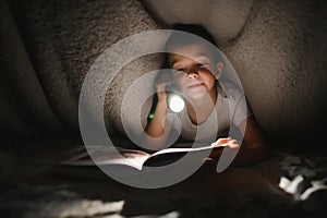 Portrait of cute little boy reading in bed with flashlight in dark room, enjoying fairytales