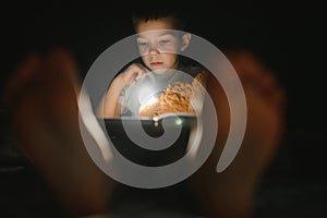 Portrait of cute little boy reading in bed with flashlight in dark room, enjoying fairytales.