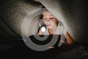 Portrait of cute little boy reading in bed with flashlight in dark room, enjoying fairytales