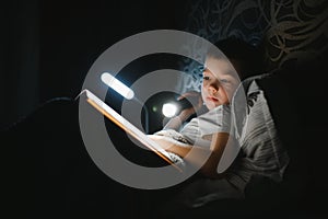 Portrait of cute little boy reading in bed with flashlight in dark room, enjoying fairytales.