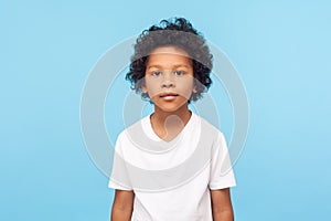 Portrait of cute little boy with stylish curly hairdo in white T-shirt standing, looking at camera with serious attentive face