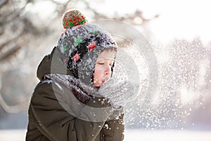 Portrait of a cute little boy in warm clothes blowing on snow outdoors during snowfall in winter sunny day