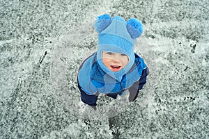 Portrait of cute little child boy in blue knitted hat on frozen pond. Winter activity outdoor. View from top