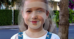 Portrait cute little child girl looking at camera standing on street in city on summer day. Young happy thinking