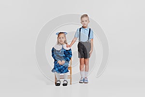 Portrait of cute little children, girl in pretty blue dress sitting on chair and boy standing near isolated over grey