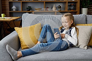 Portrait of cute little cute girl holding smartphone and resting at sofa at home. Choosing favorite music or cartoons