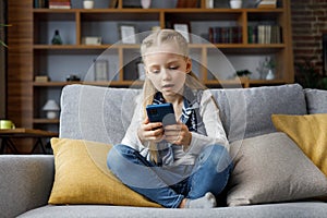 Portrait of cute little cute girl holding smartphone and resting at sofa at home. Choosing favorite music or cartoons