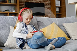 Portrait of cute little cute girl wearing earphones resting at sofa at home. Choosing favorite music or cartoons,texting