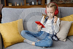 Portrait of cute little cute girl wearing earphones resting at sofa at home. Choosing favorite music or cartoons,texting