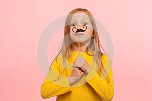 Portrait of cute little ginger girl holding funny paper mustaches and looking at camera with surprised eyes