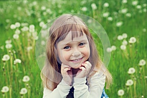 Portrait of cute little girl with beautiful smile and blue eyes sitting on the flower meadow, happy childhood concept