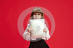 Portrait of cute little girl, child in big glasses, holding tablet against red studio background. Online school lessons