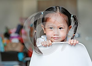 Portrait of a cute little girl dodge in the back of white plastic chair looking out for something at home