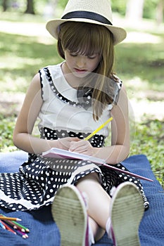 Portrait of a cute little girl in a dress with a hat while sitting on a blanket on the grass and draws in his notebook