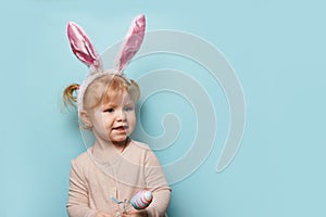 Portrait of a cute little girl dressed in Easter bunny ears holding colorful egg on blue background