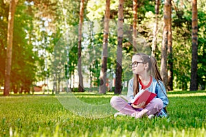 Portrait of Cute Little Girl in Glasses Sitting On Grass With Book In Park