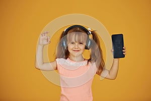 Portrait of cute little girl in headphones and with phone in the studio against yellow background