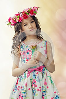 Portrait of cute little girl in nice spring dress, with flower wreath on head, holds pink rose in hands.