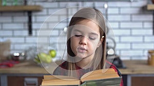 Portrait of a cute little girl in a plaid shirt reading a book sitting at the kitchen table