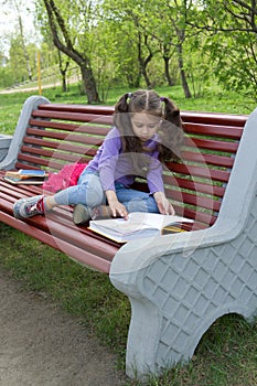 Portrait of cute little girl reading a book sitting on the wooden bench in spring city park