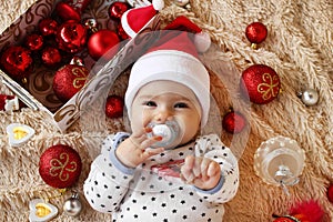 Portrait of cute little girl in Santa Claus red hat with pacifier on a beige plaid with red and white Christmas decorations.