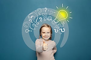 Portrait of a cute little girl showing thumb up and standing near a blue wall with a question marks and a yellow light bulb drawn