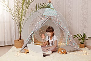 Portrait of cute little girl sitting in peetee tent and having video call or broadcasting livestream, looking at notebook screen,