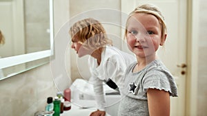Portrait of cute little girl smiling at camera while washing her face, brushing teeth together with her sibling brother