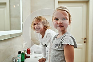 Portrait of cute little girl smiling at camera while washing her face, brushing teeth together with her sibling brother