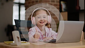 Portrait of cute little girl smiling to camera and gesturing thumb up, enjoying homeschooling via laptop and headphones