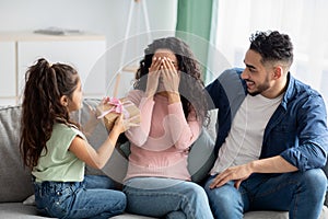 Portrait of cute little girl surprising her mom, giving present at home