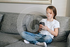 Portrait of cute little girl talking on video call by mobile phone in living room sitting on cozy sofa.