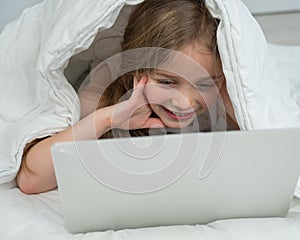 Portrait of a cute little girl using a laptop while lying under a blanket.