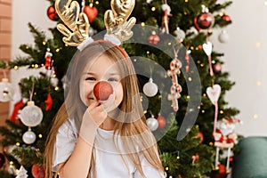 Portrait of a cute little girl wearing Rudolf horns holding a festive balloon to her nose. The child portrays a