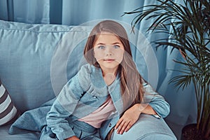 Portrait of a cute little girl wearing a stylish dress, sitting on a sofa at home, looking at a camera