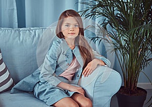 Portrait of a cute little girl wearing a stylish dress, sitting on a sofa at home, looking at a camera