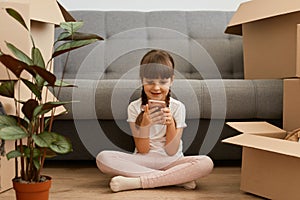 Portrait of cute little girl wearing white t shirt sitting on floor near sofa and cardboard boxes and flower, child holding cell