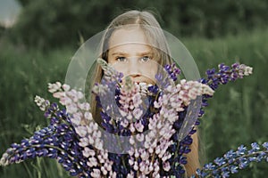 Portrait of a cute little happy seven year old kid girl with bouquet of bloom flowers lupines in a field in nature outdoor.