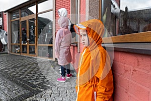 Portrait cute little lonely caucasian kid boy in bright orange waterproof raincoat stand under rain against brick wall